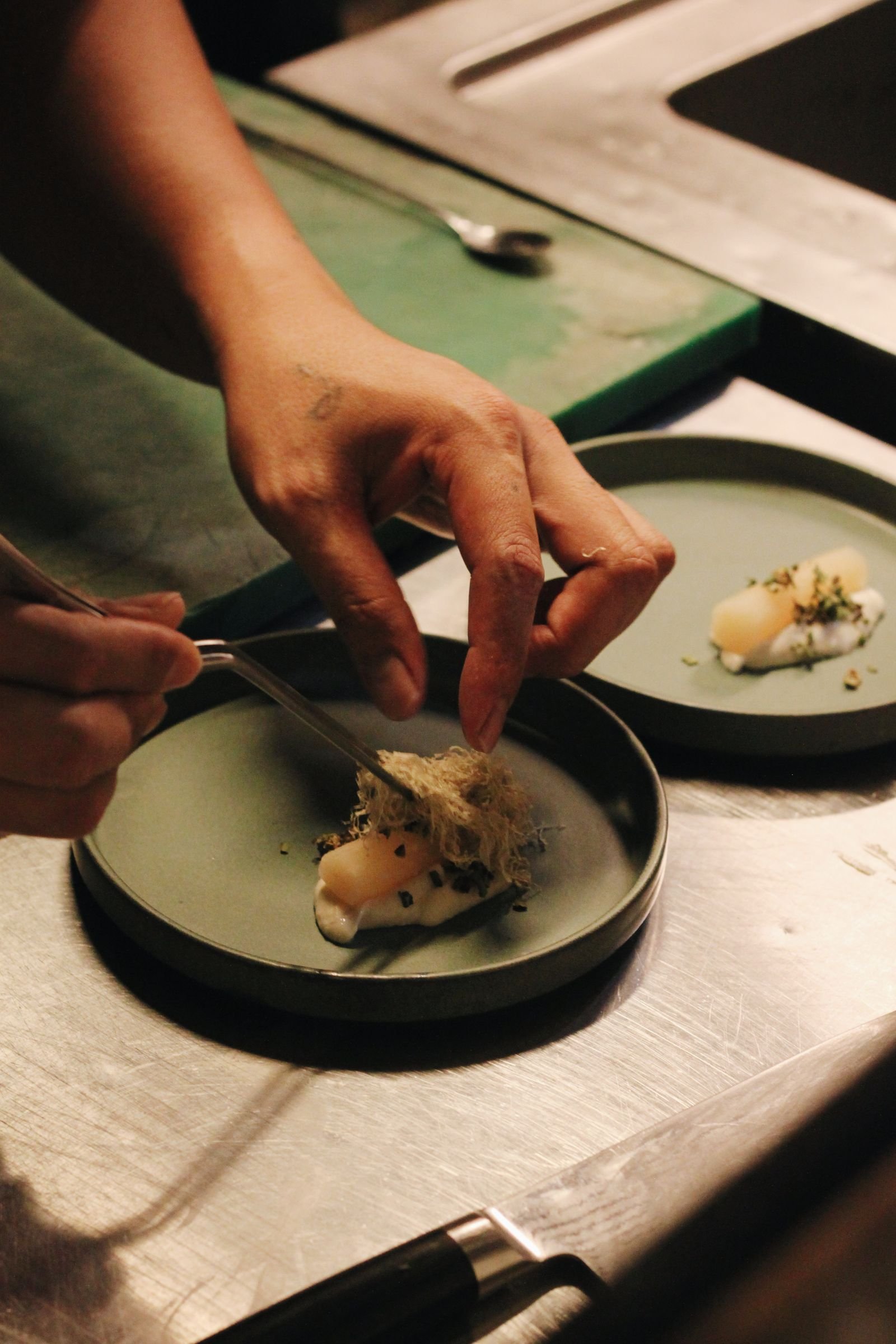 Hands plating a delicate course with tweezers in a warm, dim kitchen